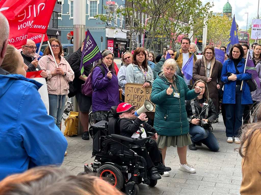 A crowd gathers at a street protest; a woman holds up a sign reading "STOP BIG SUPPLY BANK," while a person in a wheelchair uses a megaphone.